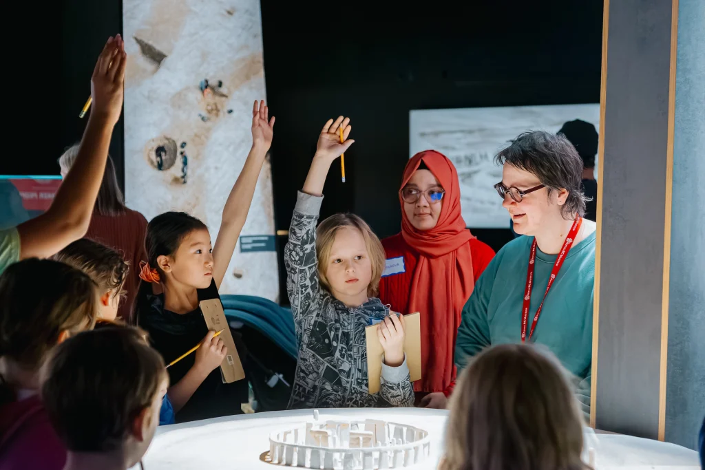 Students participating in a class activity at the museum. Many children are gathered around a white table with a small model of Stonehenge, holding papers and pencils. Three have their hands raised. On the right is a woman with grey hair and glasses, wearing a red lanyard and speaking to them. Behind, a woman wearing glasses and a red hijab looks on thoughtfully.