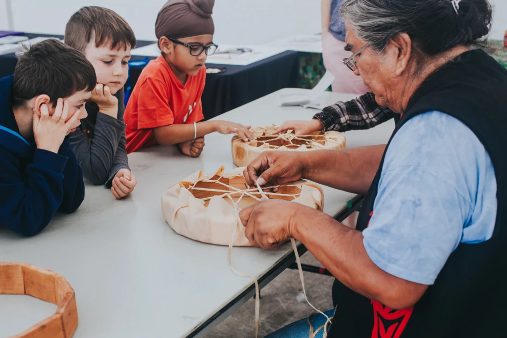Artist Virgil Sampson sitting at a table, tying the strings to a leather-hide drum. He has grey hair, glasses and is wearing a black vest with red formline designs. Two boys sit across from him looking on. To the right, a boy, wearing glasses and a patka, touches another drum being held out by another person out of view.