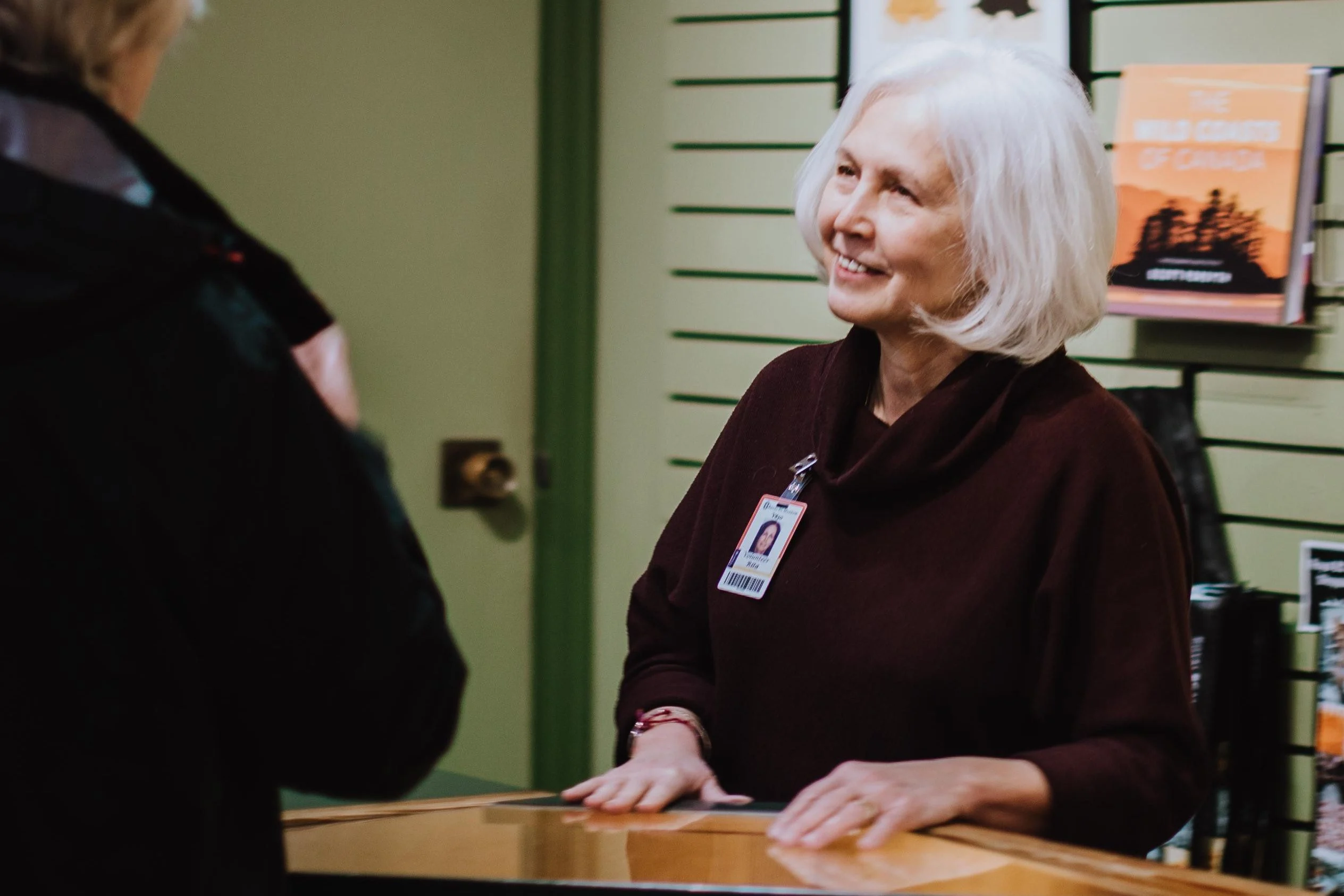 A museum volunteer smiling at an information desk. She has a white bob and is wearing a badge pinned to her sweater. Behind her is a green slated wall with books on display. On the left, a visitor is out of focus talking to her.