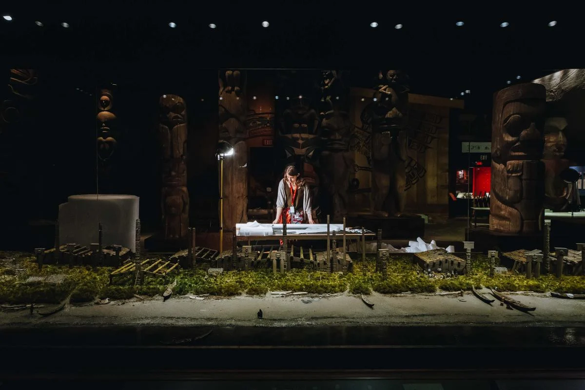 Wide shot of a museum worker standing in a gallery, inspecting papers on a table. She is illuminated by a bright lamp, while the rest of the gallery is dimly lit in dramatic shadow. In the background are many tall, carved wooden poles in a Northwest Coast Indigenous style. In the foreground is a miniature recreation of a seaside Indigenous village featuring carved poles, longhouses and house posts amid trees. A sandy beach with several canoes completes the scene.