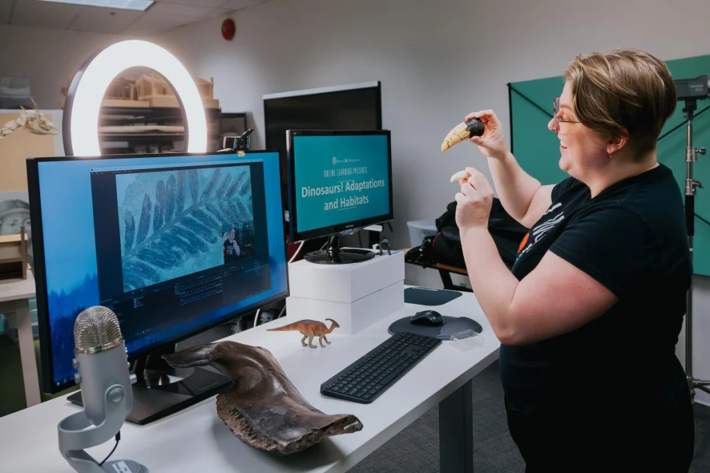 Museum staff member leading a digital field trip. She stands at a desk outfitted with a video streaming setup: ring light, microphone and multiple screens that show video feed and presentation deck. She is smiling at a webcam, holding two large tooth fossils. On the desk is also a large fossil fragment and small dinosaur figurine.