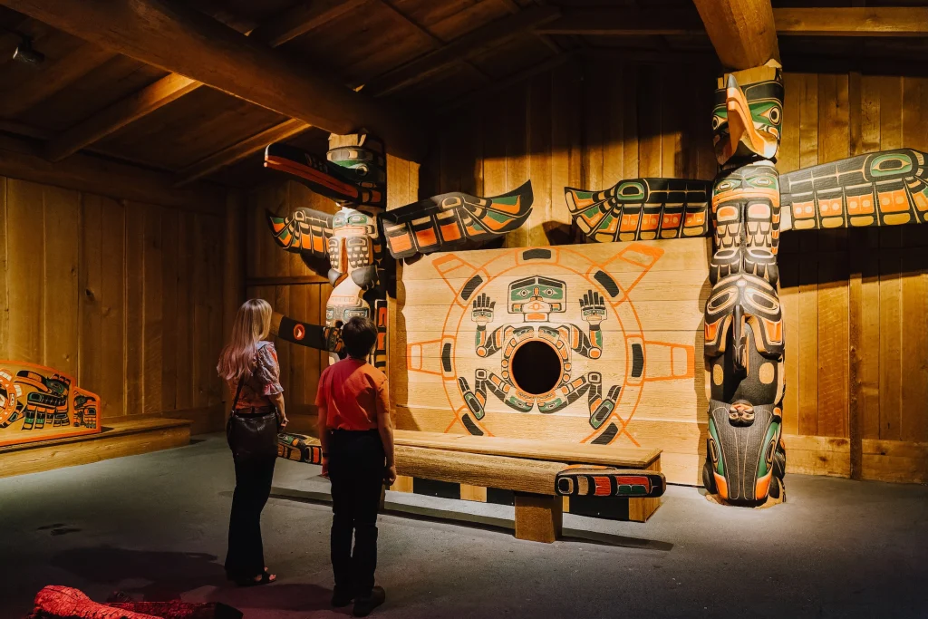 Interior of Jonathan Hunt House being admired by two museum visitors. The installation has two large carved wooden poles flanking a large rectangular painted panel. The left pole is of a raven figure and the right pole is of an eagle. Both have outstretched wings and are carved in Northwest Coast formline style and painted black, red, and green. The panel in the middle has a sun design with a human figure, and a circular hole in the torso. A wooden platform and bench sit in front. The room’s walls are panelled wood, and there is an A-frame pointed roof. A painted bench against the left wall is halfway in frame.