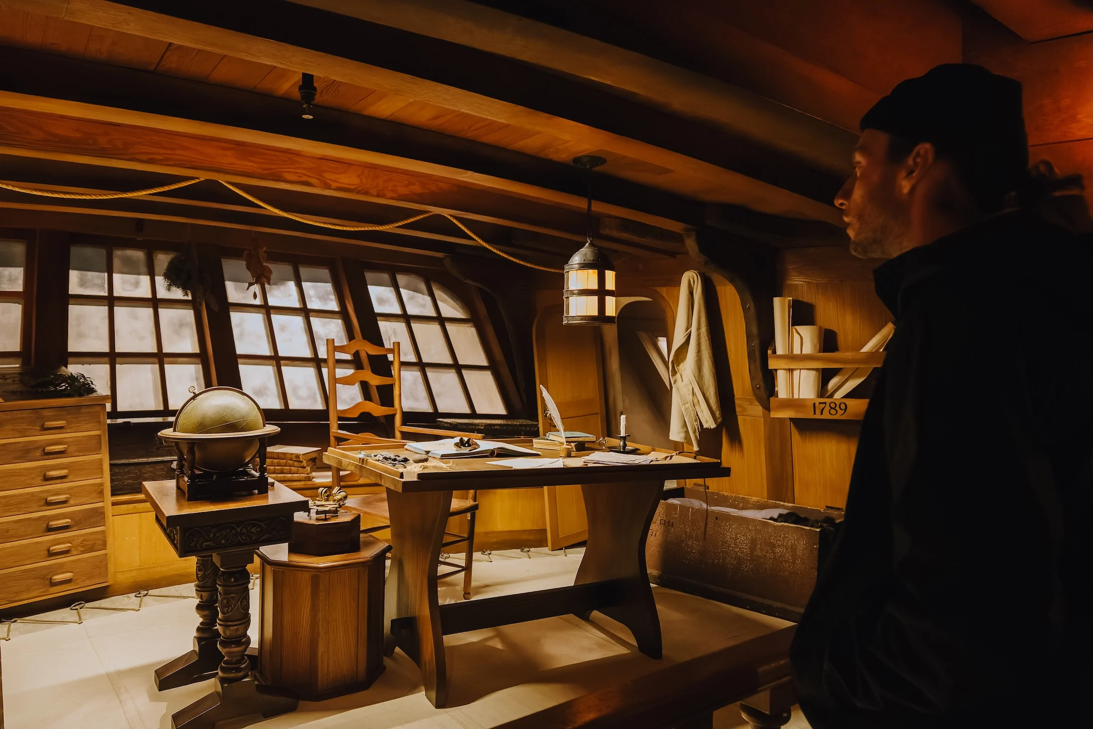 A visitor inside of the HMS Discovery exhibition, a full-scale replica of a wooden boat from the late 1700s. They are looking at the boat’s interior, where there is a desk, chair and several side tables. On display are a globe, several navigation instruments, quill and inkwell, and books spread about. In the background are shelves installed into the walls and a set of panelled windows.