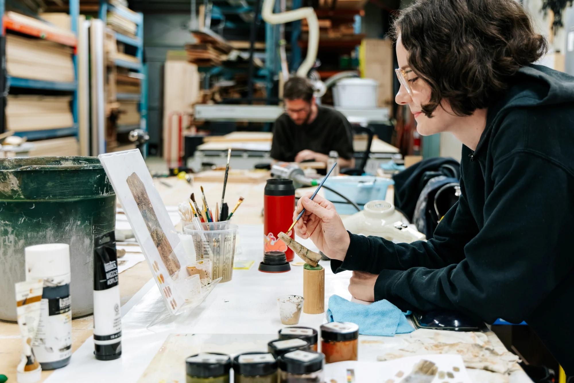 A museum worker seated at a table, painting a bone replica in a fabrication studio. On the table is a reference image, supplies for painting and an open coffee tumbler. In the background is another museum worker leaning over the table and tall, ceiling-high industrial shelves with wood planks. The image hums with creative energy.