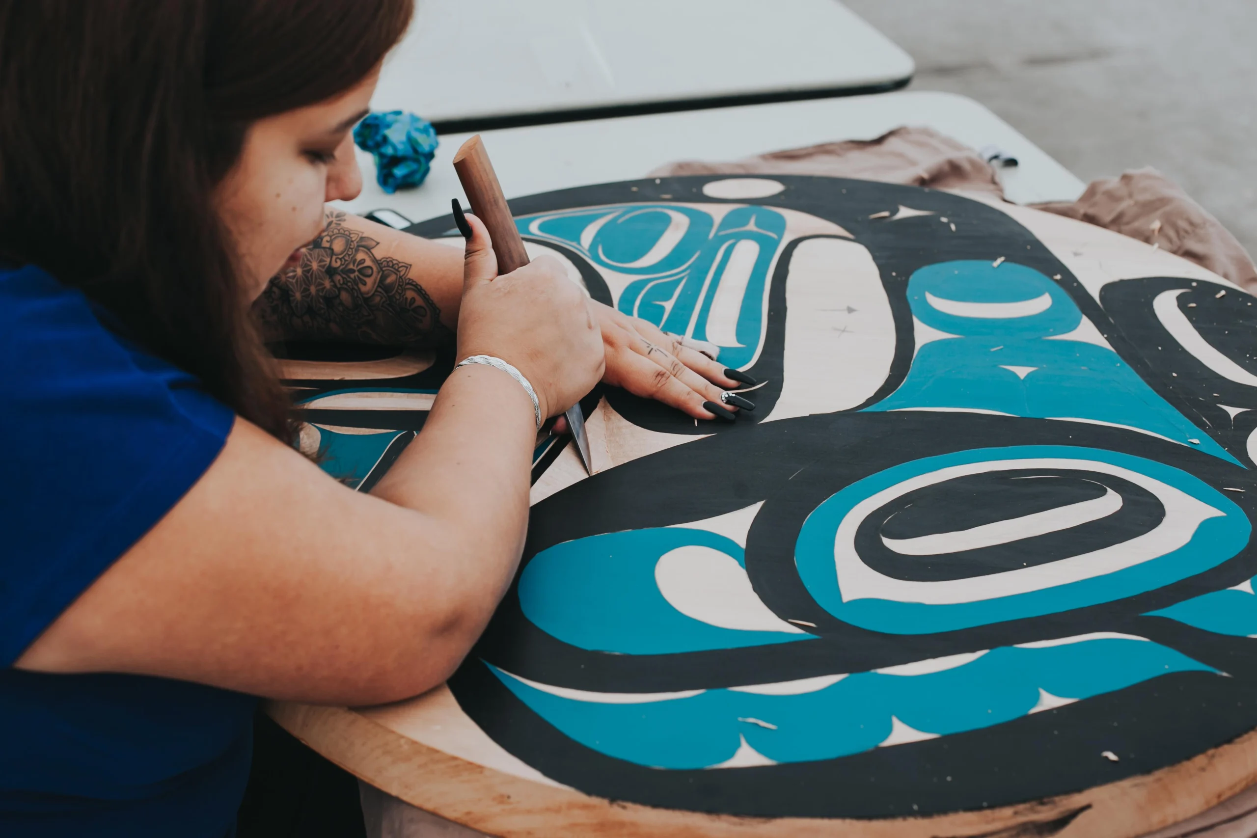 Closeup of artist Joslyn Williams working on a painted wooden carving. We see Joslyn from the side, holding a knife and carving on a large circular piece of wood that has a formline whale painted on it in black and turquoise. Joslyn’s tattooed arm with long fingernails rests atop the carving.