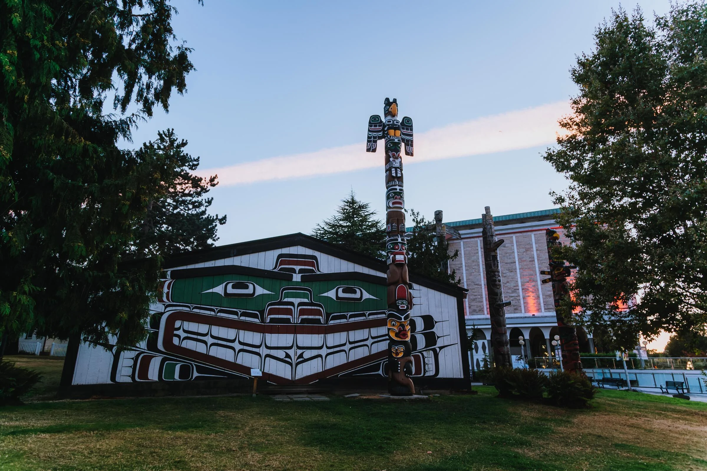 Wide shot of Mungo Martin House, Wawadiťła, at dusk. The house has a white painted exterior with a Great Killer Whale Northwest Coast formline design in black, green and red. A heraldic carved wooden pole stands in front with yellow, black, red, green and white paintings of animal figures including an eagle at the top. In the background are several other wooden poles, tall trees, and the Royal BC Museum building.
