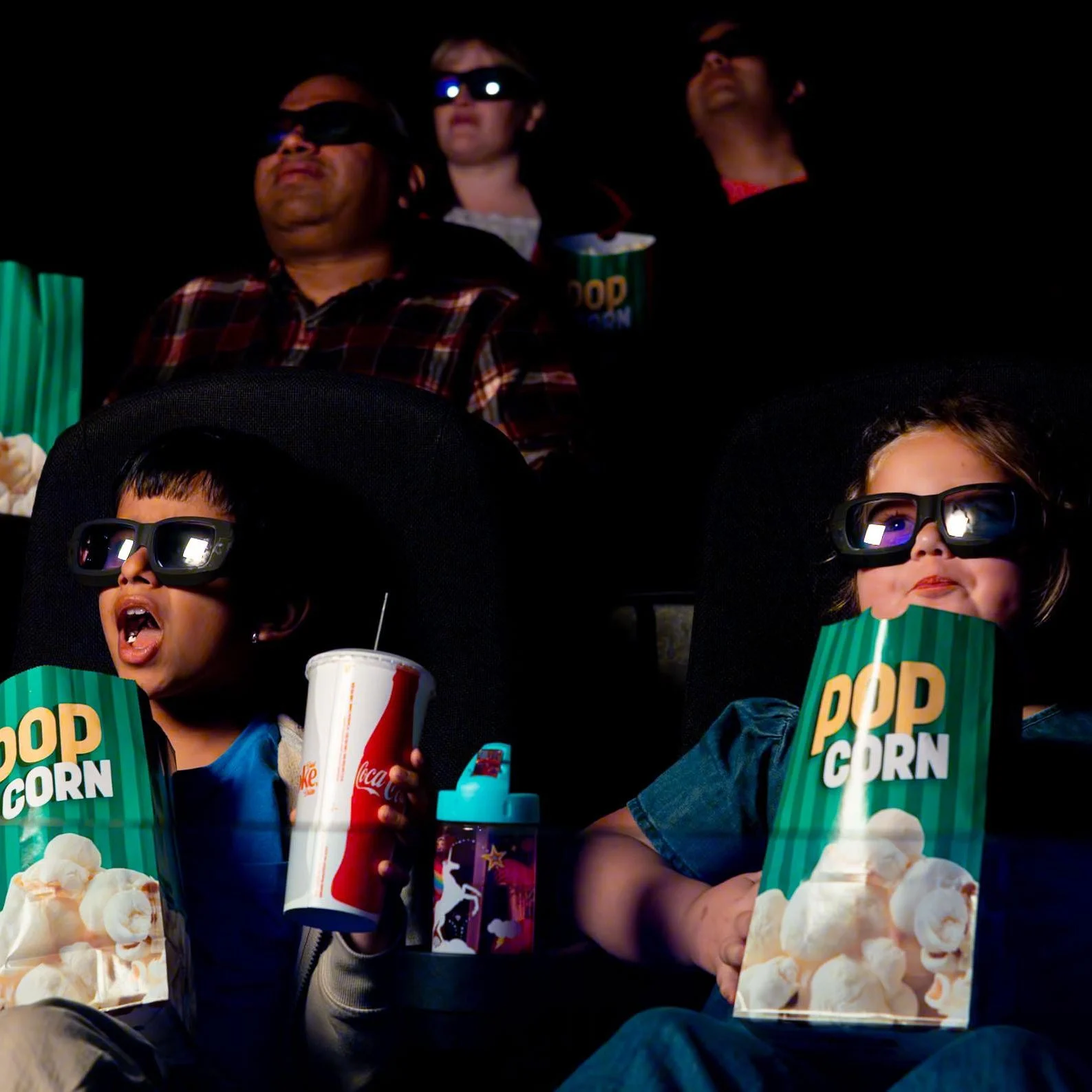Visitors sitting inside a darkened movie theatre. In the foreground on the left is a boy with dark hair wearing 3D glasses, holding a popcorn and soft drink with mouth agape. Beside is a girl, also wearing 3D glasses and holding popcorn, smiling. Behind them are other adults out of focus wearing their own 3D glasses and popcorn.