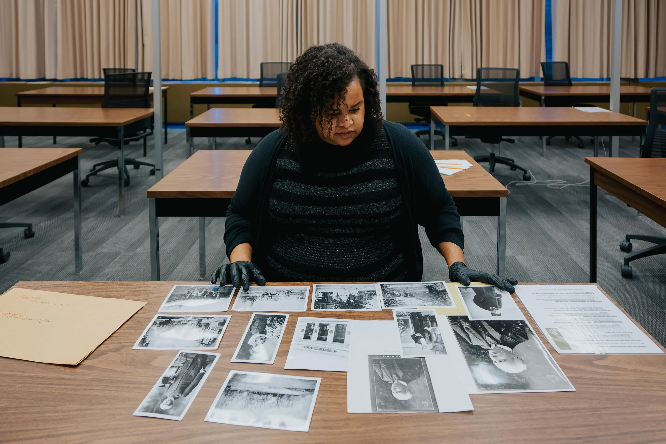 A woman with curly black hair sits at a table. She is wearing black disposable gloves and examining a table full of black and white photographs. Behind her are many more similar tables sitting empty inside the BC Archives building.