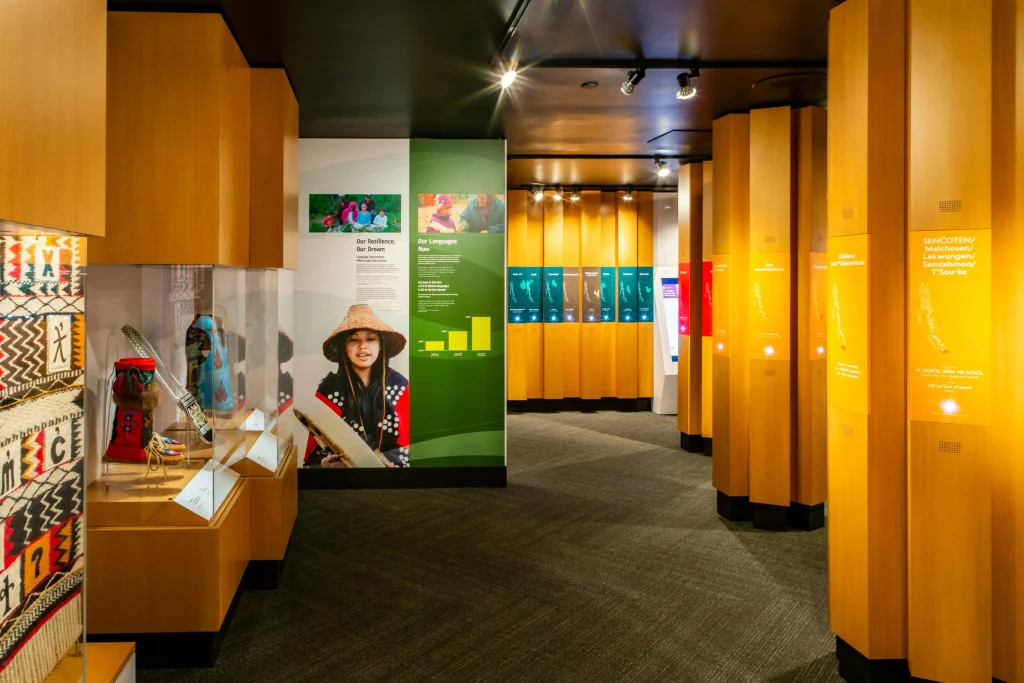 Wide shot of the Our Living Languages exhibition. Tall wooden panelled displays span the height of the room. To the left is a colourful Salish weaving, and glass displays featuring beaded leather hide boots, a purse and embellished vest. Facing the camera is an informational wall display showing a person wearing a pointed cedar hat and button blanket who is playing a leather drum, with supporting text and graphs. On the far back and right walls are beam-like displays that show maps of different languages in British Columbia and have speakers and illuminated buttons.