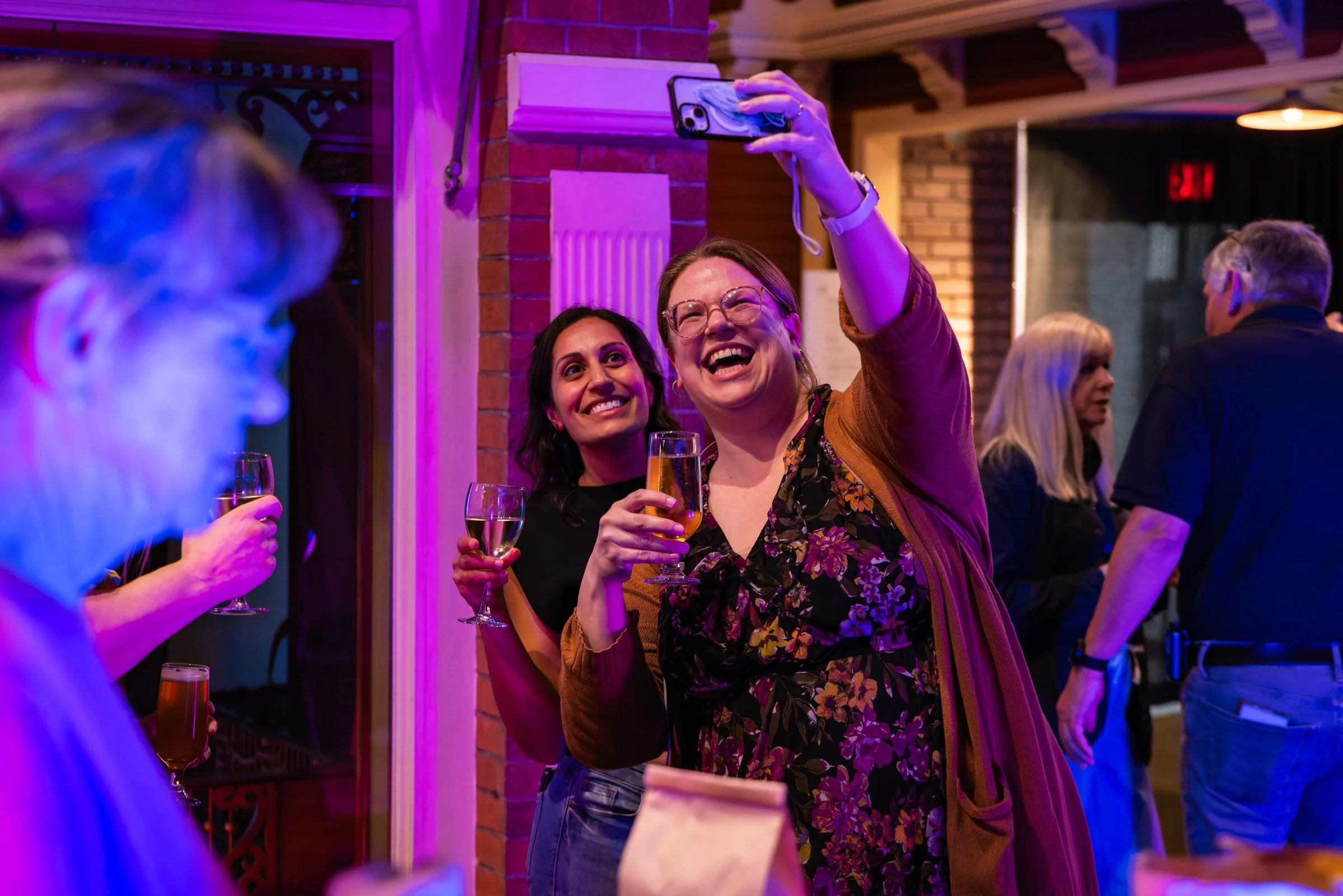 Wide shot of two women taking a smiling selfie inside the musem. The woman taking the picture in front has brown hair and glasses. She holding a glass of beer and is wearing a floral dress and orange cardigan. The woman behind has black hair and is holding a glass of white wine and wearing a black shirt. Behind them is a brick wall. They are bathed in pink and blue light, and around them are other museum guests milling about.
