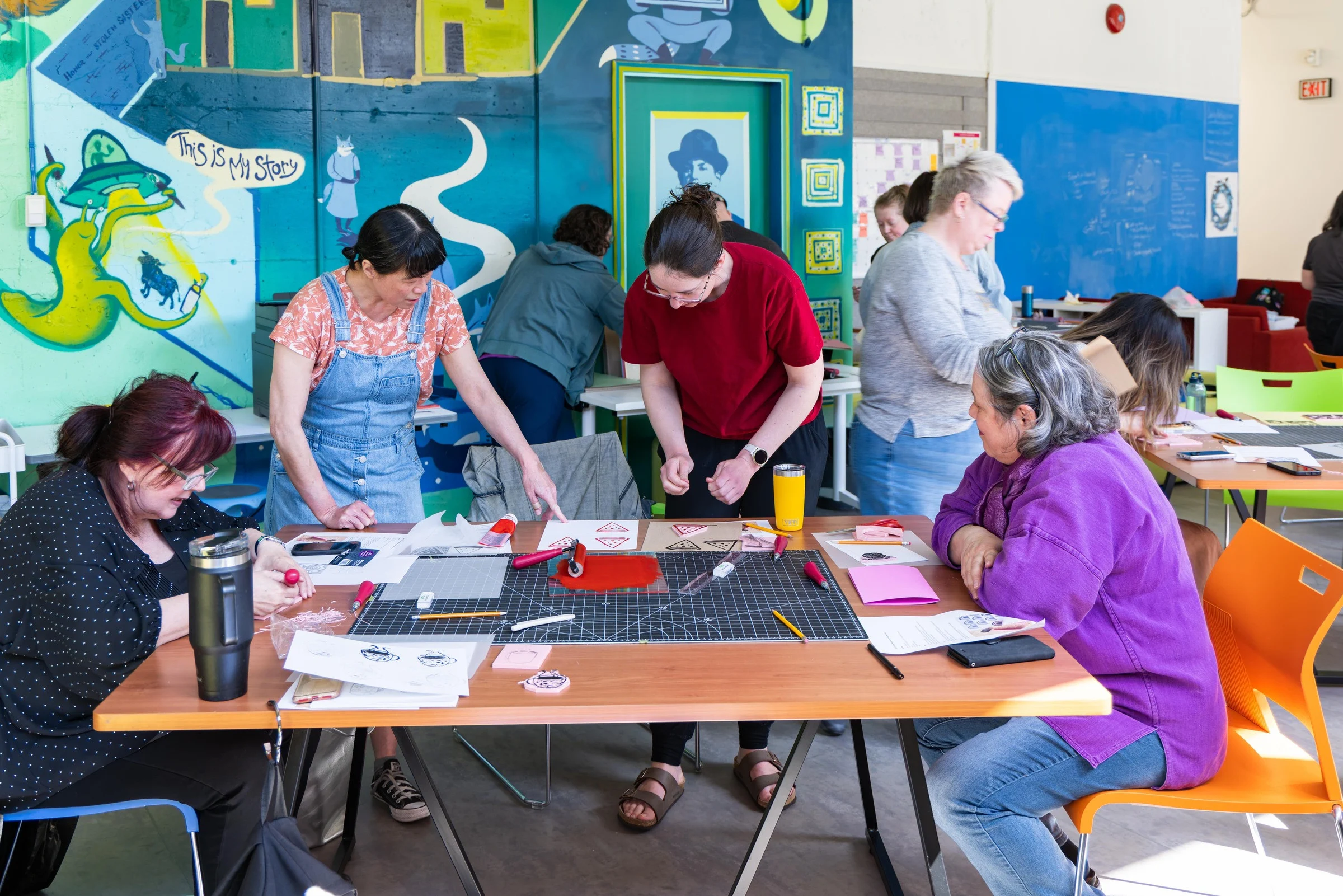 Artist Marlene Yuen hosting a workshop. She is wearing blue overalls over a floral t-shirt, and is pointing at papers on a table. Another participant stands beside her in red. They are flanked by two more seated participants, each with their own papers.The table is strewn with a cutting mat and paper-cutting supplies. In the background are similar tables and a large turquoise and green painted mural. The entire room has a colourful, creative energy.