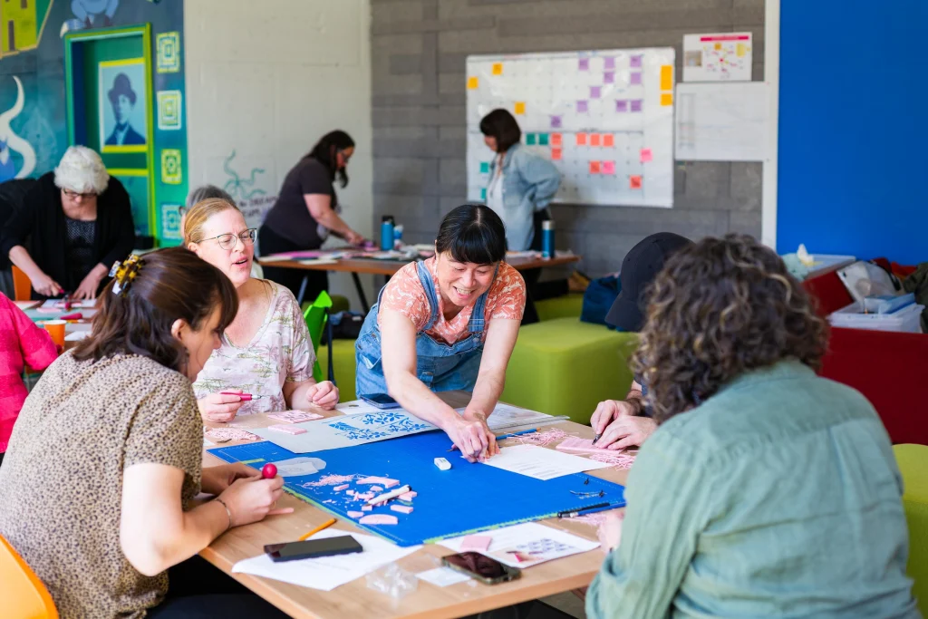Artist Marlene Yuen hosting a workshop. She is wearing blue overalls over a floral t-shirt, and is leaning over a table filled with cutting mats, printing blocks and paper. Several other participants seated at the table are carving their own blocks. In the background are similar craft tables, green ottoman seats, a turquoise mural and a calendar with sticky notes. The entire room has a colourful, creative energy.