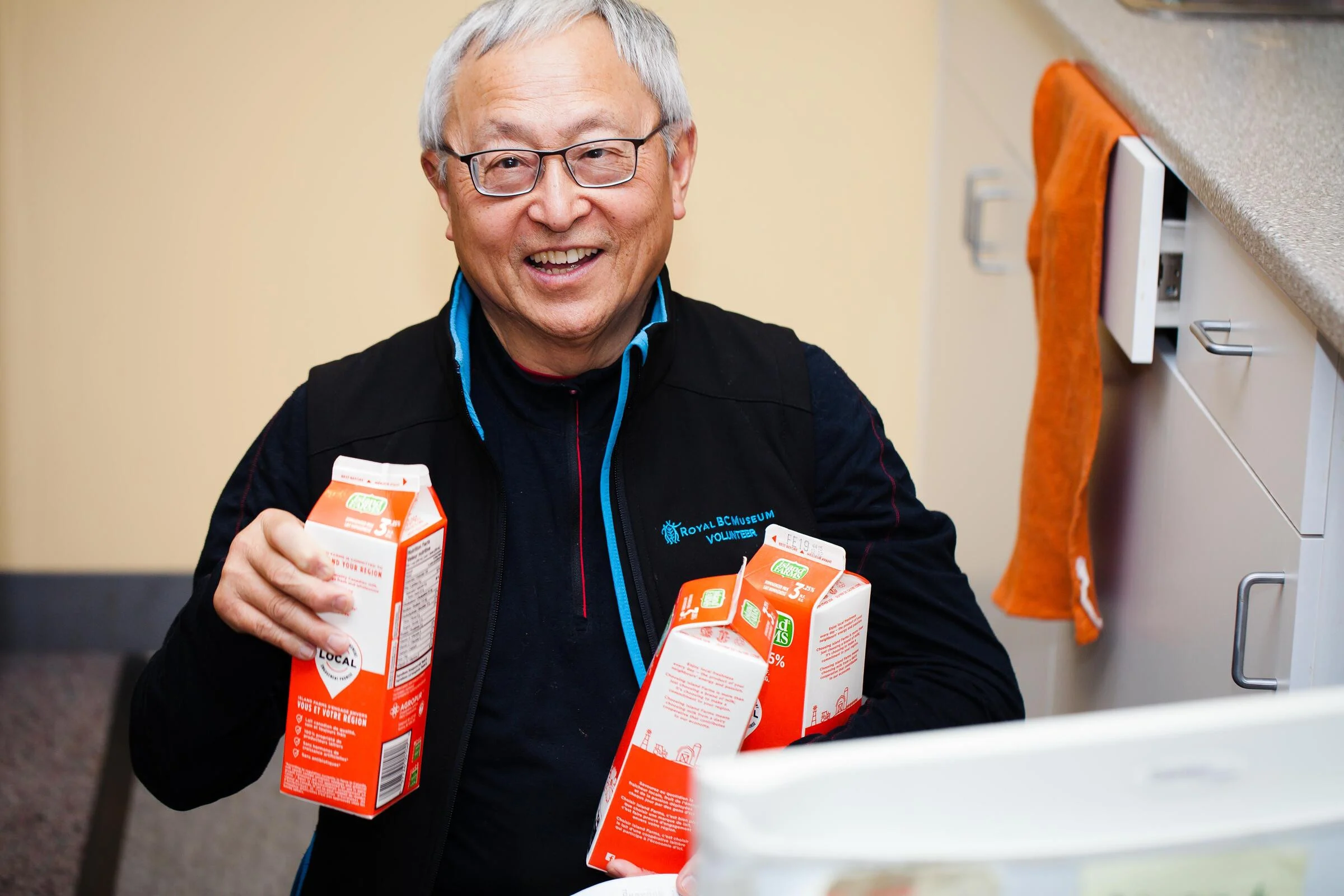 A man with grey hair and glasses, holding three cartons of milk. He is smiling and wearing a black volunteer vest. He is in a kitchen and the open fridge door is just peeking into the frame.