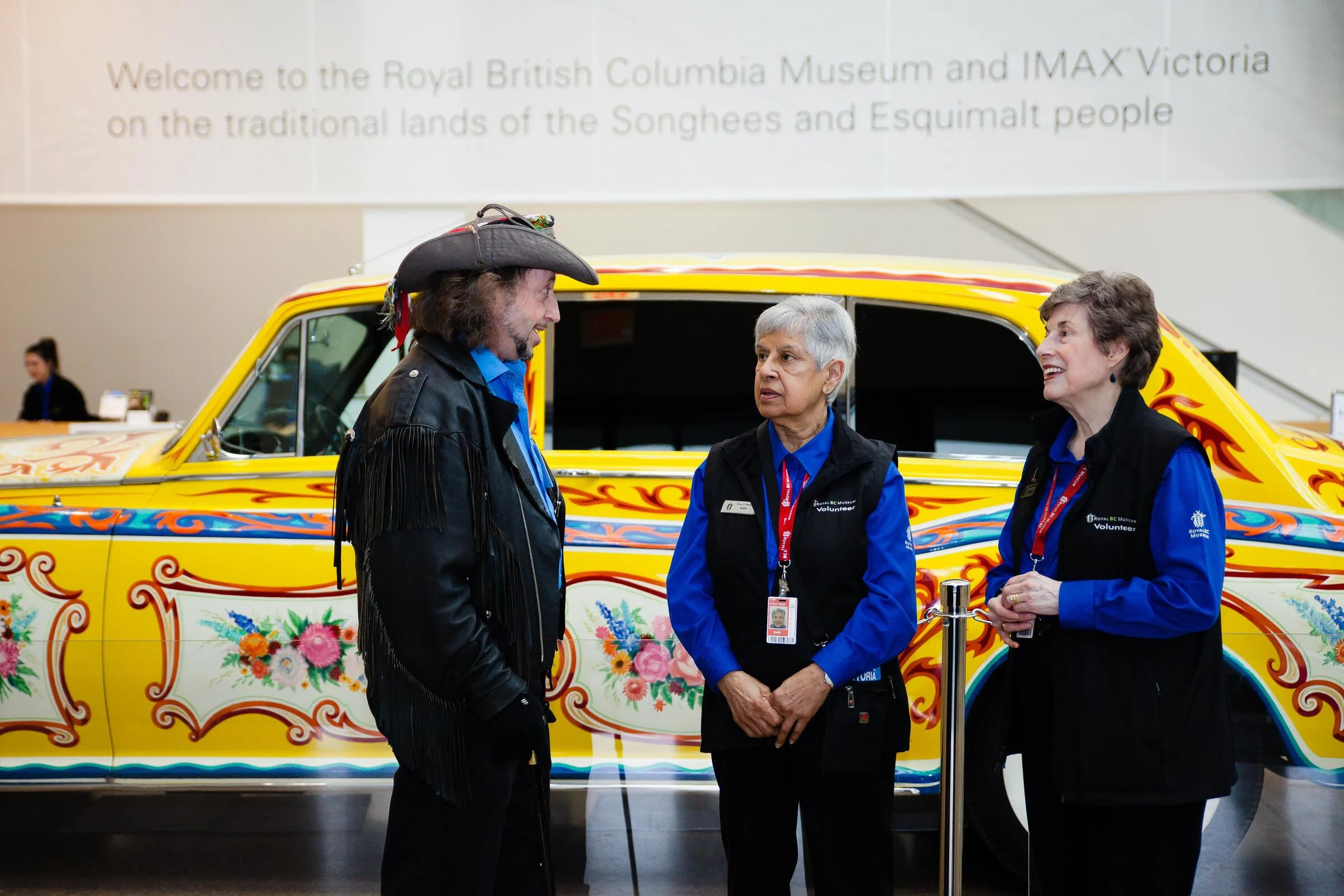 Three people talking in the museum lobby. On the left, a man wearing pirate hat and fringed leather jacket speaks animatedly. The other two volunteers are women with short hair, wearing uniforms of red lanyard and black vests over blue shirts. Behind them is John Lennon’s Psychedelic Rolls-Royce, a yellow car with curlicue and floral painted details all over. Above them is a banner that says “Welcome to the Royal British Columbia Museum and IMAX Victoria on the traditional lands of the Songhees and Esquimalt people.”